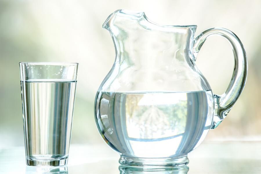 A clear pitcher and glass filled with water. It is shot straight on on a glass table, with a nature background seen through a window. It has a narrow depth of field.